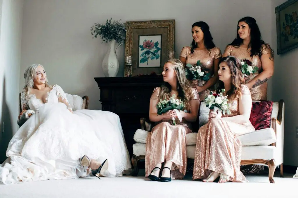 A bride in a lace dress and bridesmaids in sequin gowns share a candid moment indoors.