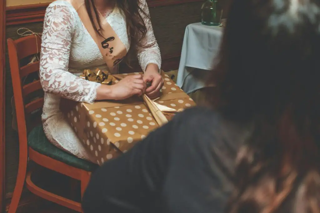 A woman at a bridal shower unwrapping a polka dot gift indoors, wearing a sash.